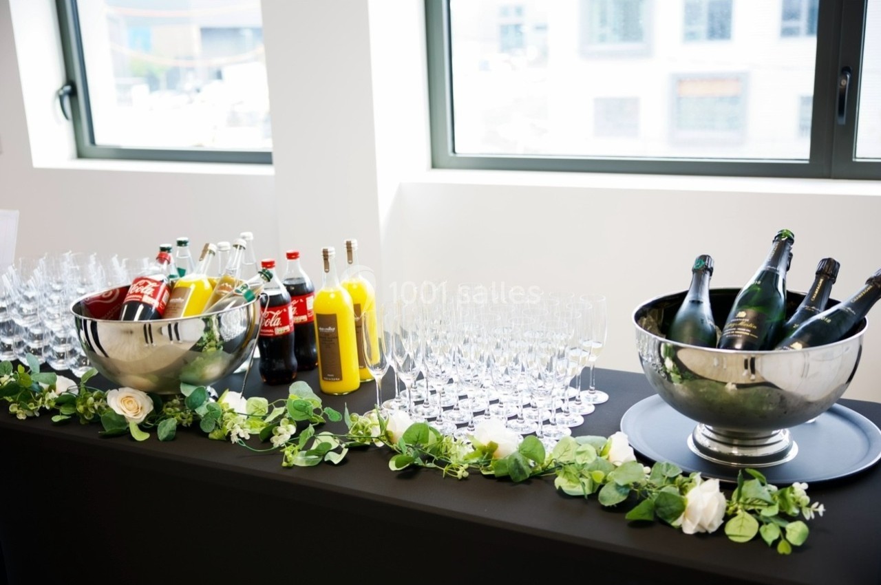 Table avec boissons variées, bouteilles de champagne, sodas et jus, entourées de verres et décorée de feuillage.