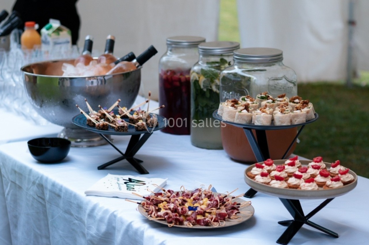 Buffet avec amuse-bouches variés, boissons en carafes et seau à glace sur une table blanche en extérieur.
