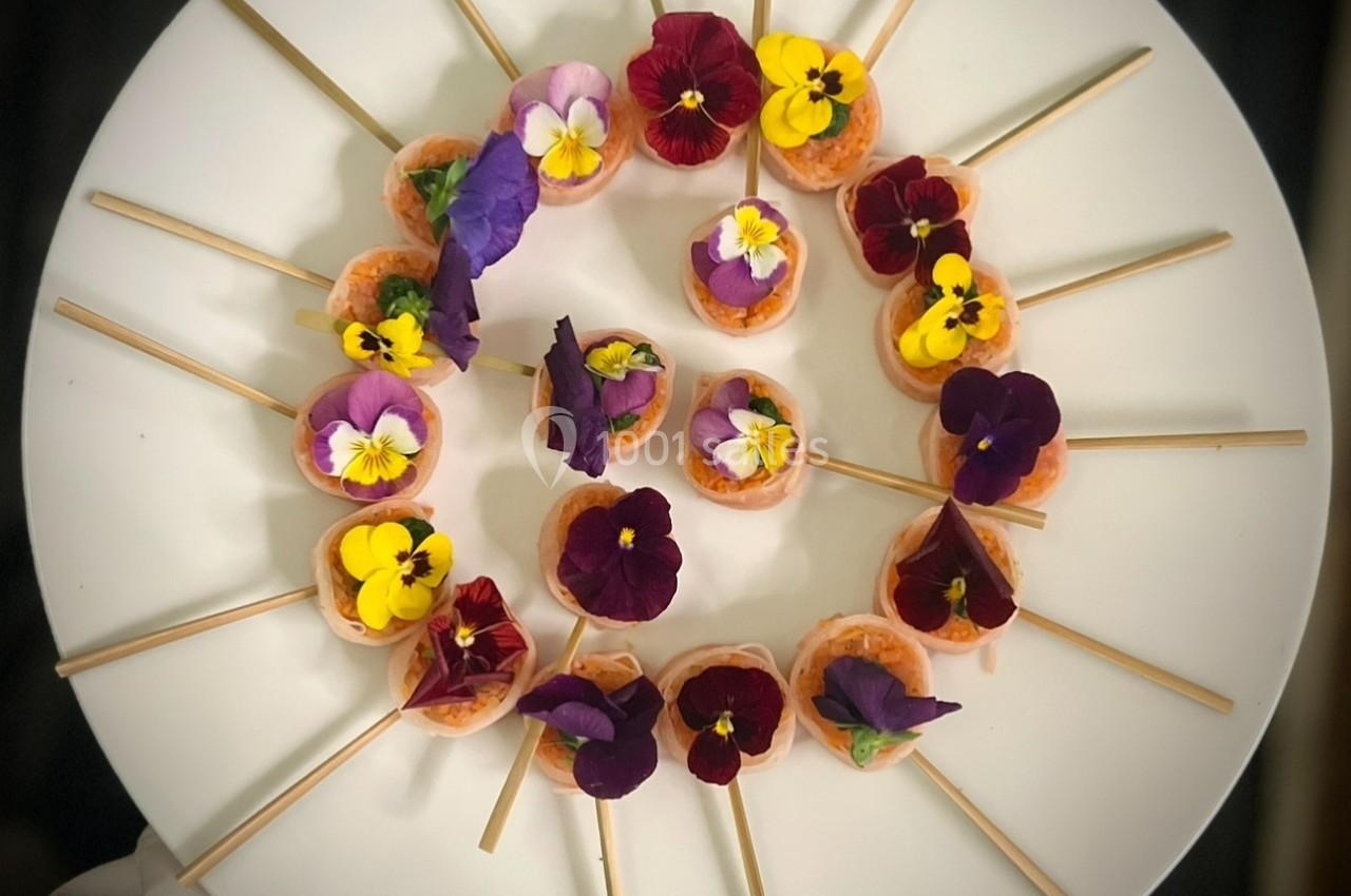 Assortiment de bouchées décorées de fleurs comestibles colorées, disposées en cercle sur une assiette blanche.