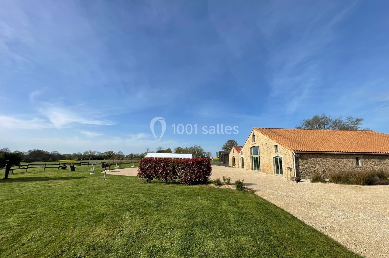 Bâtiment en pierre avec toit en tuiles, entouré de pelouse, d'arbustes et d'un ciel dégagé.