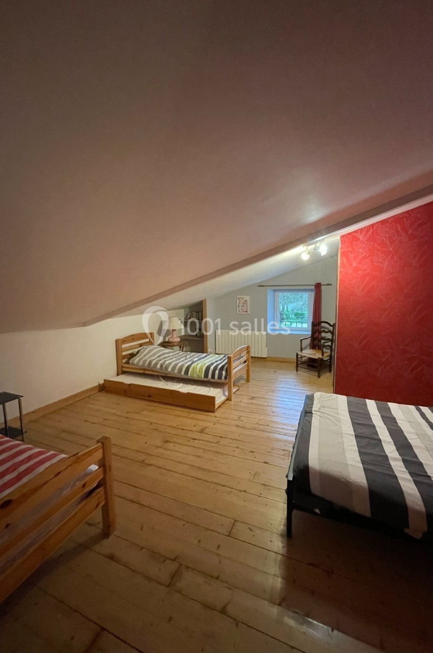 Chambre mansardée avec trois lits simples, parquet en bois, mur rouge décoratif et fenêtre donnant sur l'extérieur.