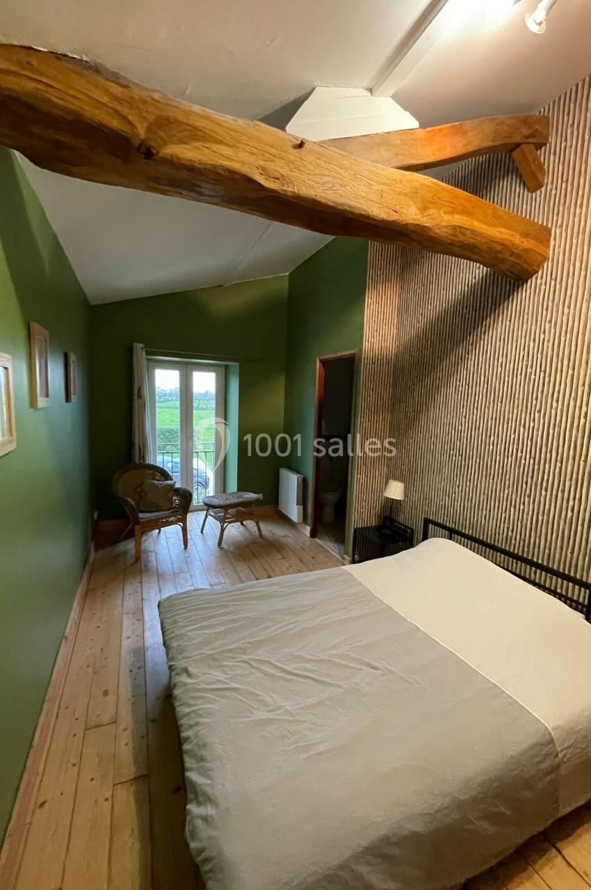 Chambre avec murs verts, lit double, parquet en bois, poutre apparente et vue sur un paysage extérieur par une porte-fenêtre.