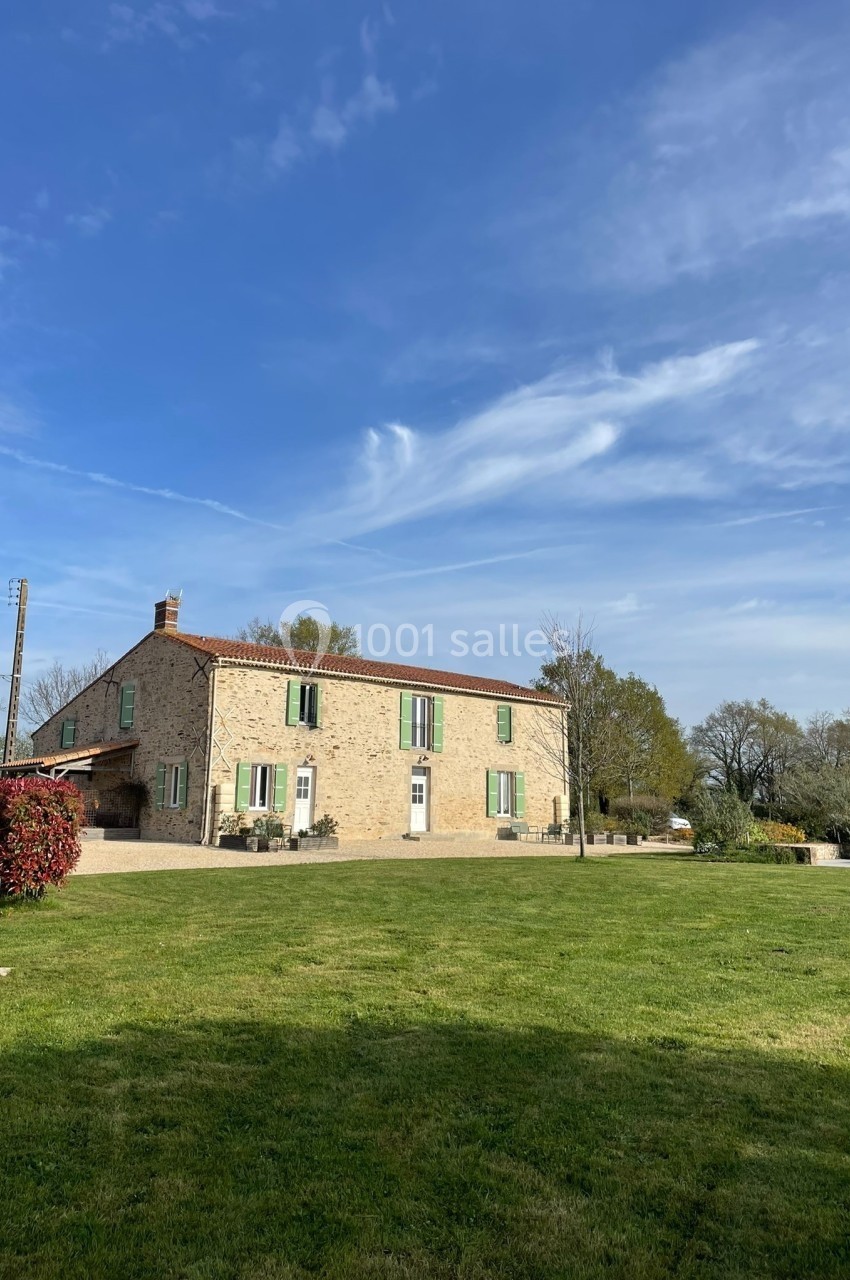 Grande maison en pierre avec volets verts, entourée d'un jardin verdoyant sous un ciel bleu dégagé.