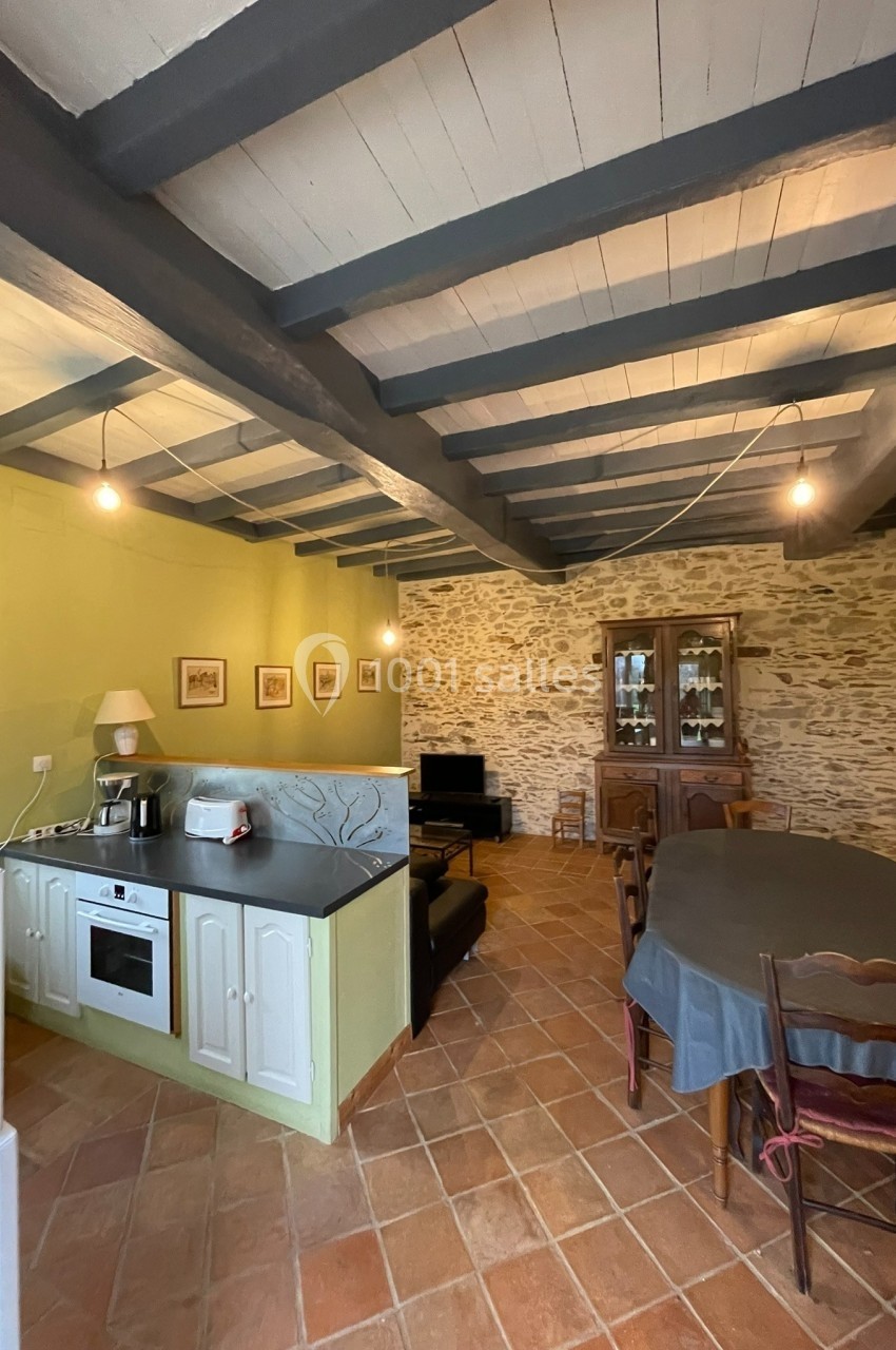 Cuisine ouverte avec murs en pierre, poutres apparentes, table à manger, et meubles rustiques sur un sol en terre cuite.