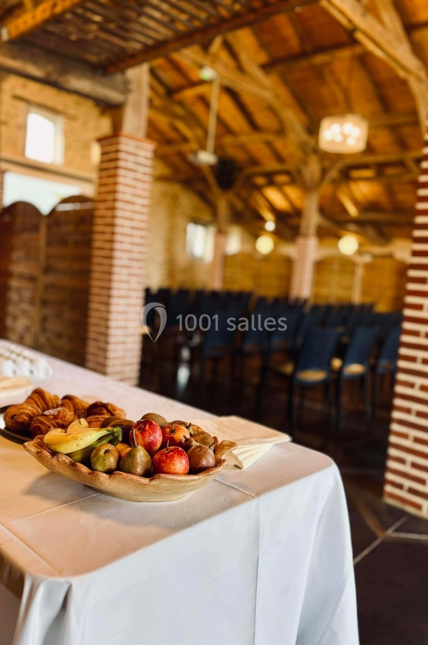 Plateau de fruits et viennoiseries sur une table blanche dans une salle rustique avec des chaises alignées en arrière-plan.