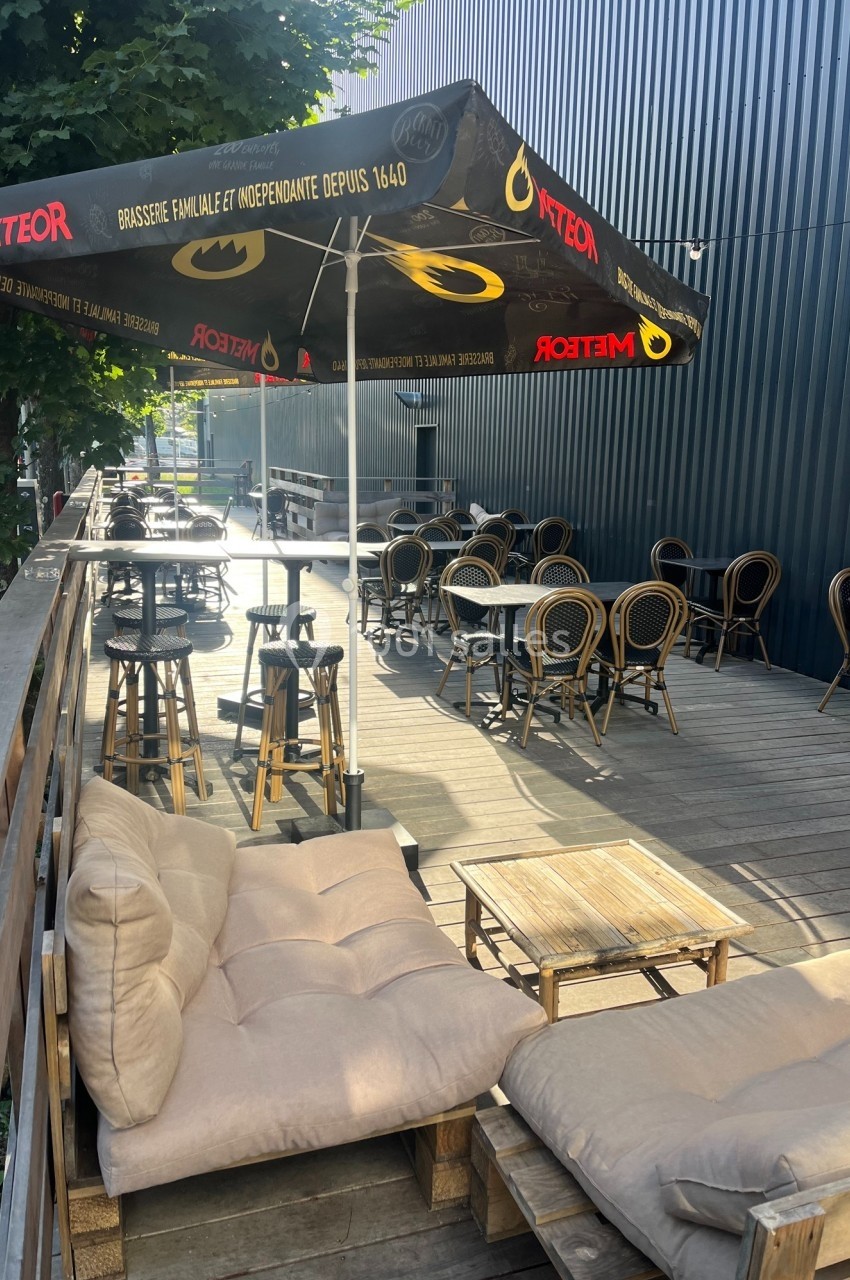 Terrasse en bois avec tables, chaises en rotin et parasols noirs, entourée de verdure et d'un bâtiment sombre.