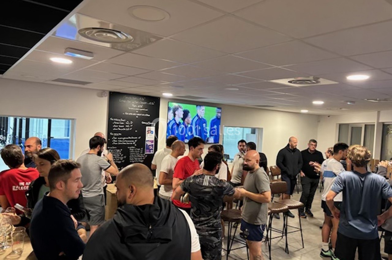 Groupe de personnes discutant dans une salle lumineuse avec un écran diffusant un match en arrière-plan.