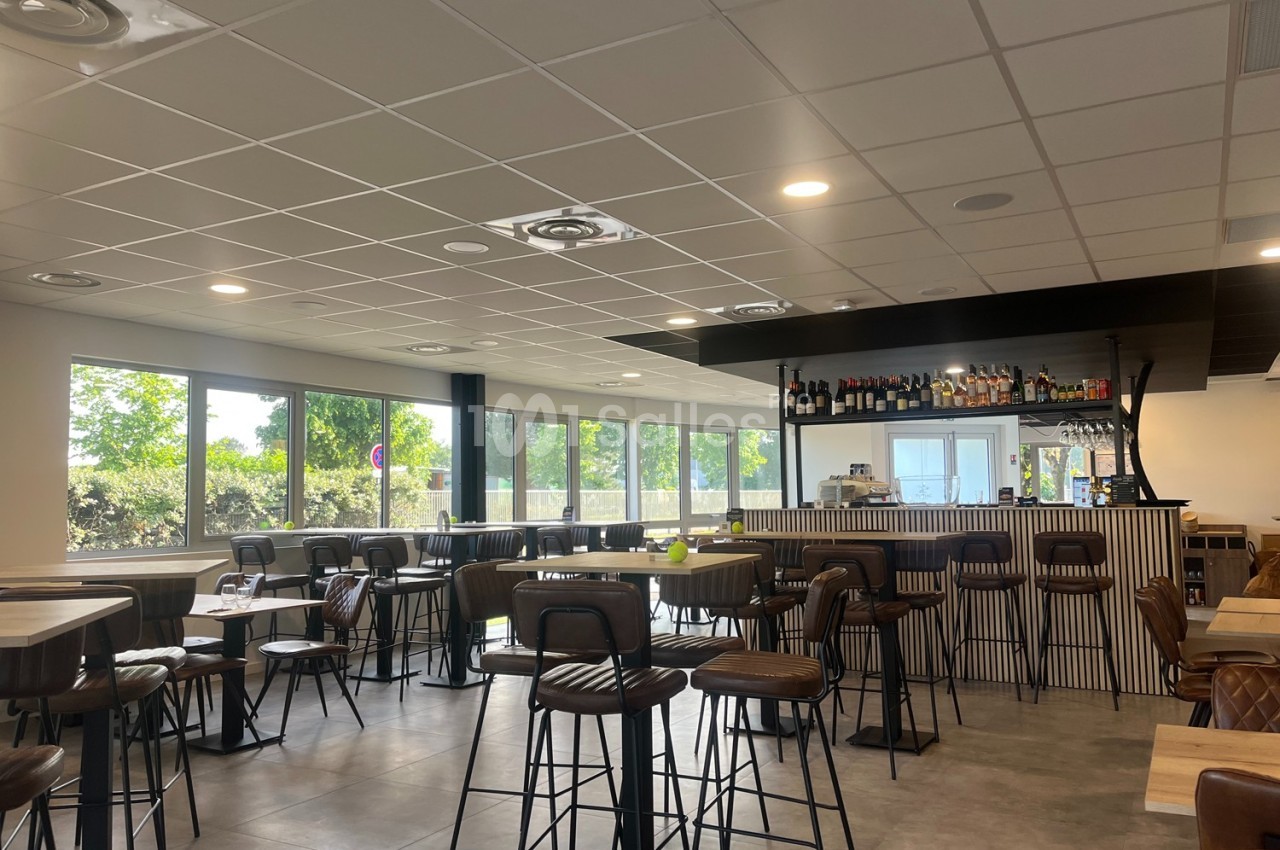 Salle de restaurant lumineuse avec un bar central, des tables hautes et des chaises en cuir, vue sur l'extérieur.