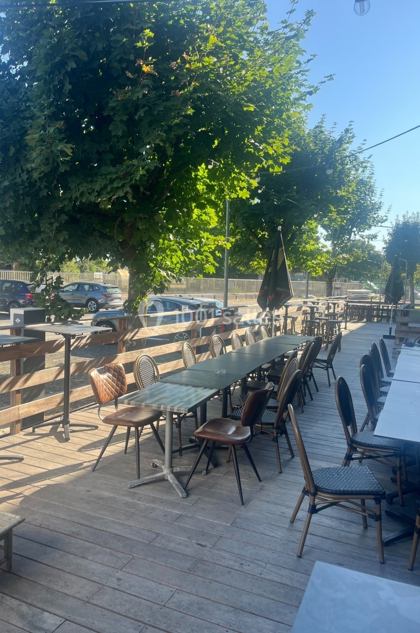 Terrasse en bois avec tables et chaises vides, ombragée par des arbres, située près d'une route et d'un parking.