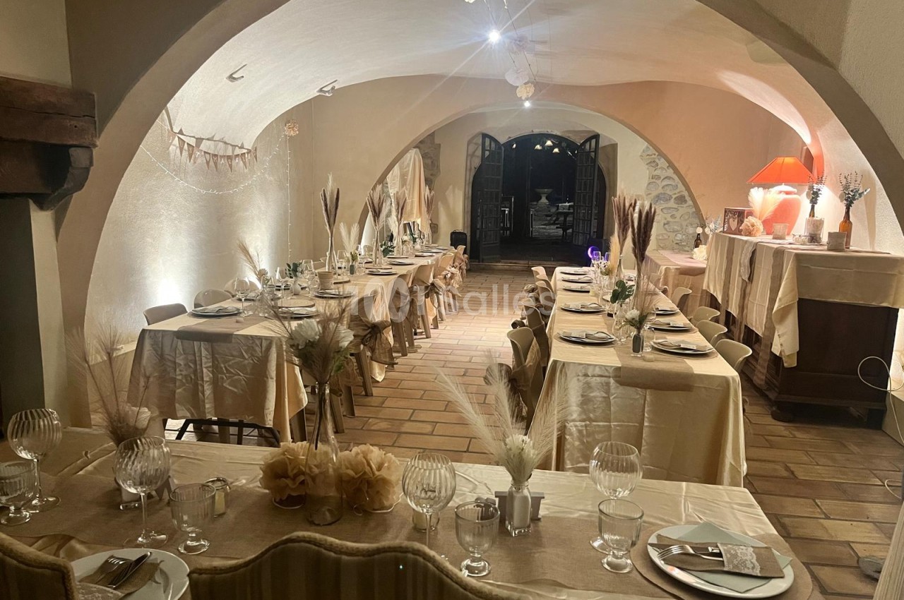 Salle voûtée en pierre dressée pour un banquet, avec plusieurs longues tables habillées de nappes beige, de roseaux secs et de verres à pied.