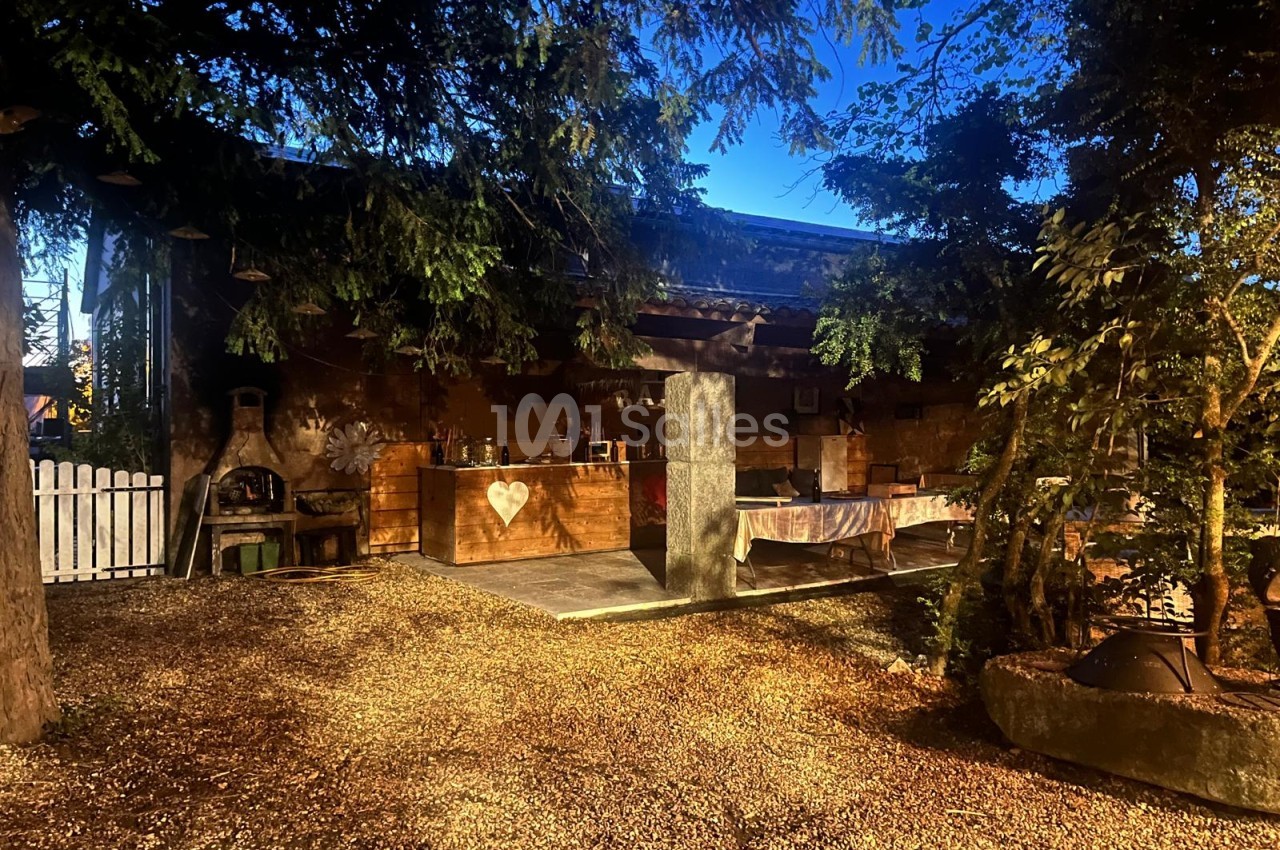 Cour extérieure de nuit sous les arbres, avec un bar en bois orné d'un cœur, une cheminée en pierre à gauche et une table dressée à nappe claire.