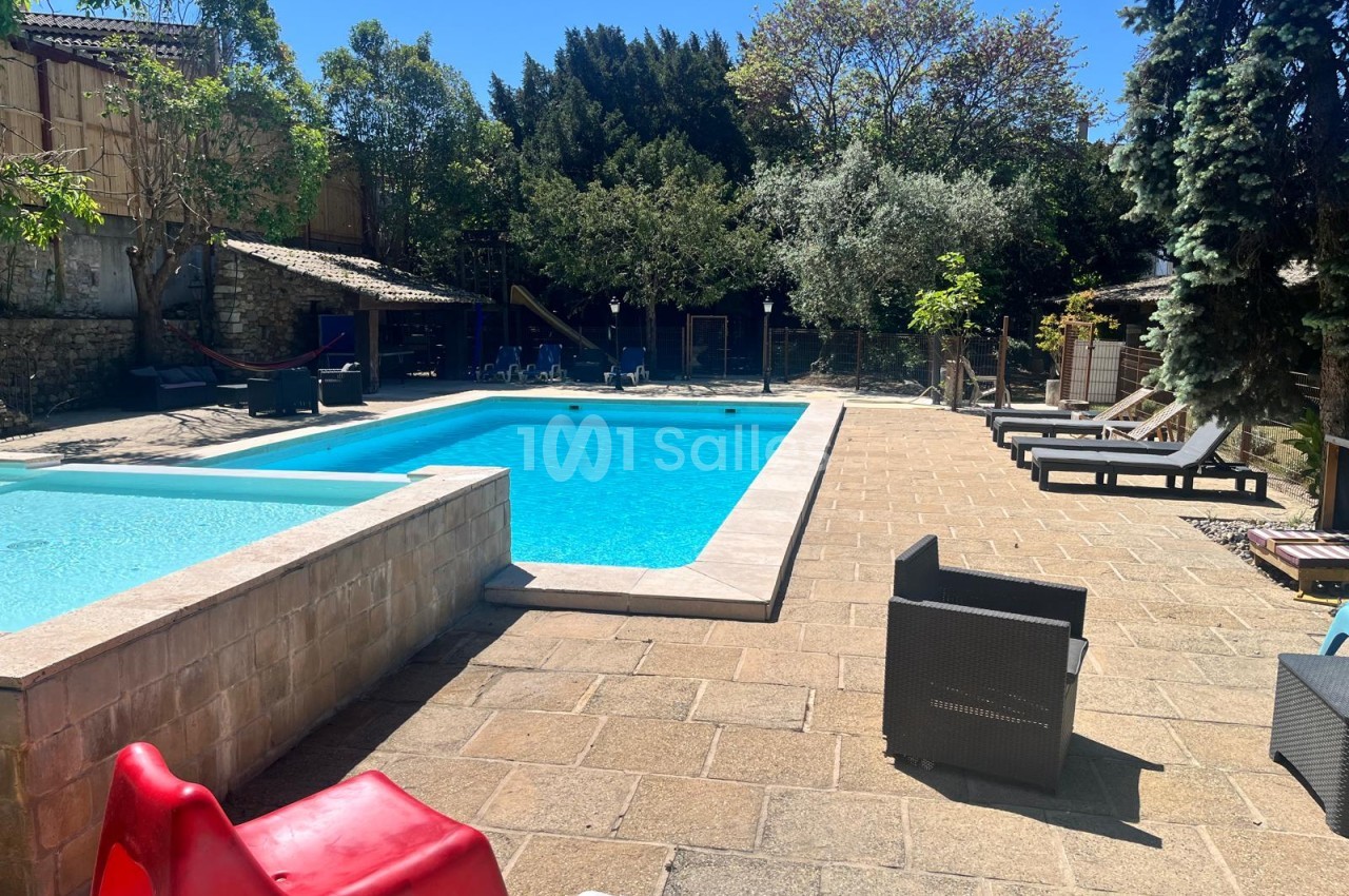 Piscine extérieure rectangulaire bordée d'une dalle en pierre, avec transats et fauteuils en résine sous des arbres, par une journée ensoleillée.