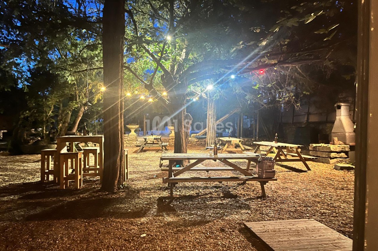 Espace extérieur de nuit sous les arbres, avec tables de pique-nique en bois, tabourets hauts et guirlandes lumineuses suspendues.