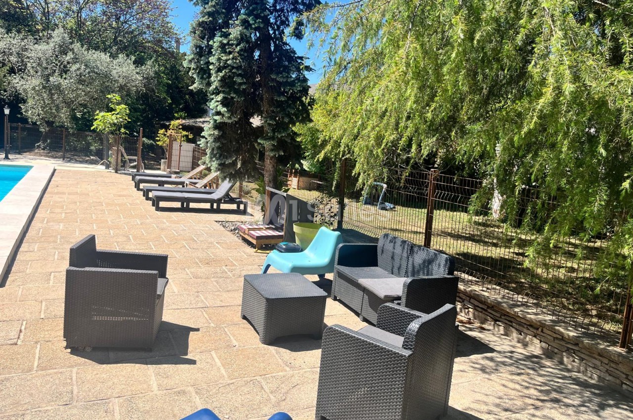 Terrasse en pierre à côté d'une piscine, avec des fauteuils et une table en résine tressée grise, des bains de soleil alignés et des arbres sous un ciel bleu.