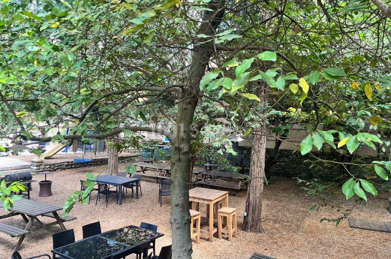 Espace extérieur ombragé sous de grands arbres avec tables et chaises noires, tables en bois hautes et bancs de pique-nique sur un sol de gravillons, aire de jeux en arrière-plan.