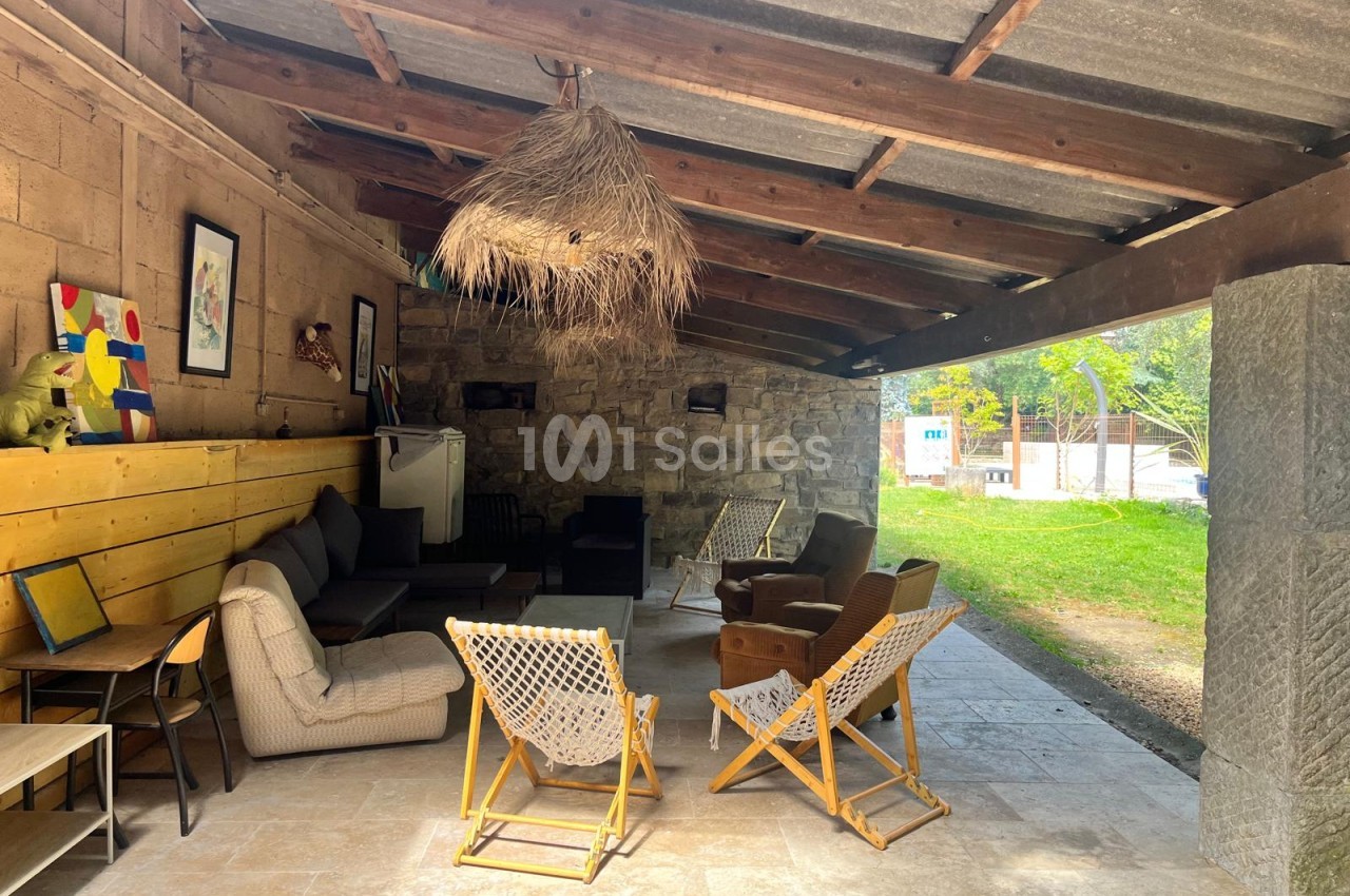 Espace couvert et ouvert sur jardin, meublé de canapés, fauteuils et chaises longues en toile, avec une suspension en paille et un mur en pierre apparente.