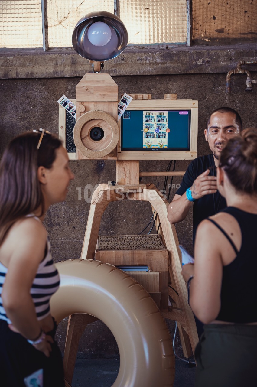 Une borne photo en bois avec écran et accessoires, entourée de personnes discutant dans un espace industriel.