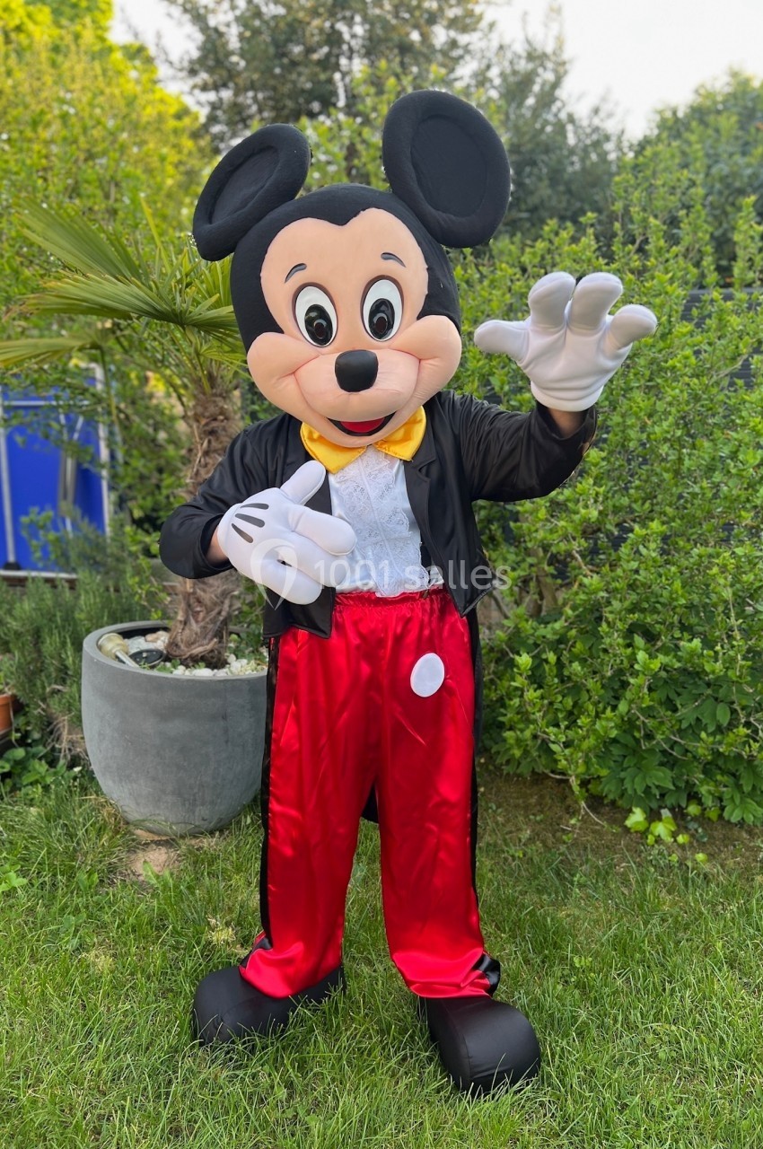 Personne déguisée en personnage de souris anthropomorphe, posant dans un jardin avec des plantes en arrière-plan.