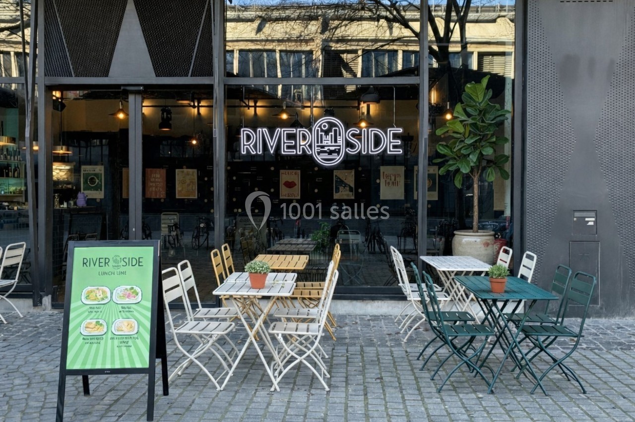 Terrasse d'un café-restaurant avec des tables et chaises en métal, devant une vitrine affichant le nom ’Riverside’.