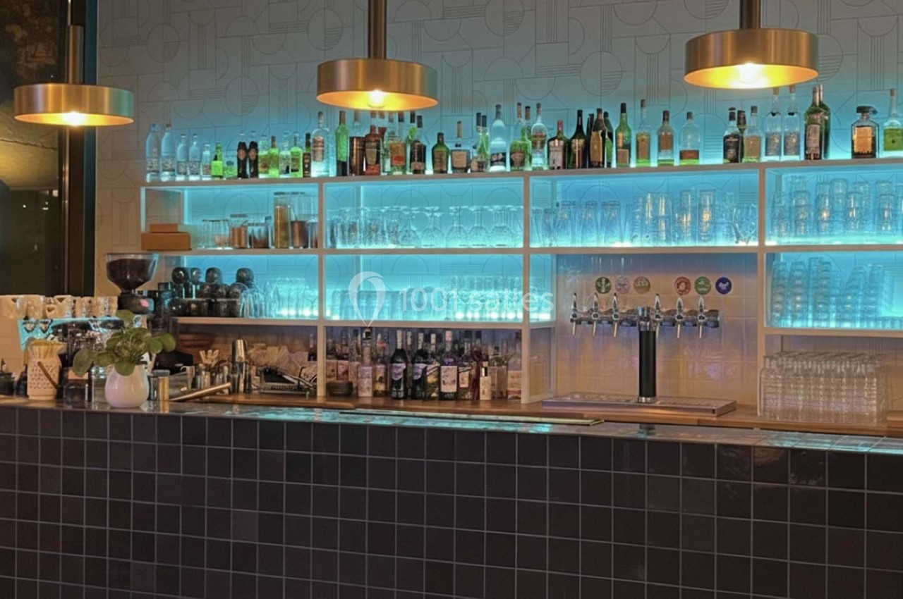 Bar moderne avec étagères éclairées affichant des bouteilles, verres et tireuses à bière, sous des lampes suspendues.