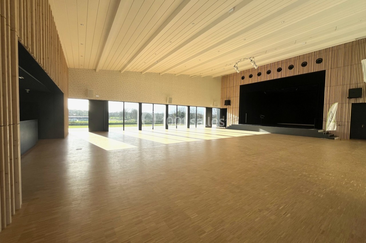 Salle spacieuse avec parquet clair, grandes baies vitrées laissant entrer la lumière et une scène au fond.