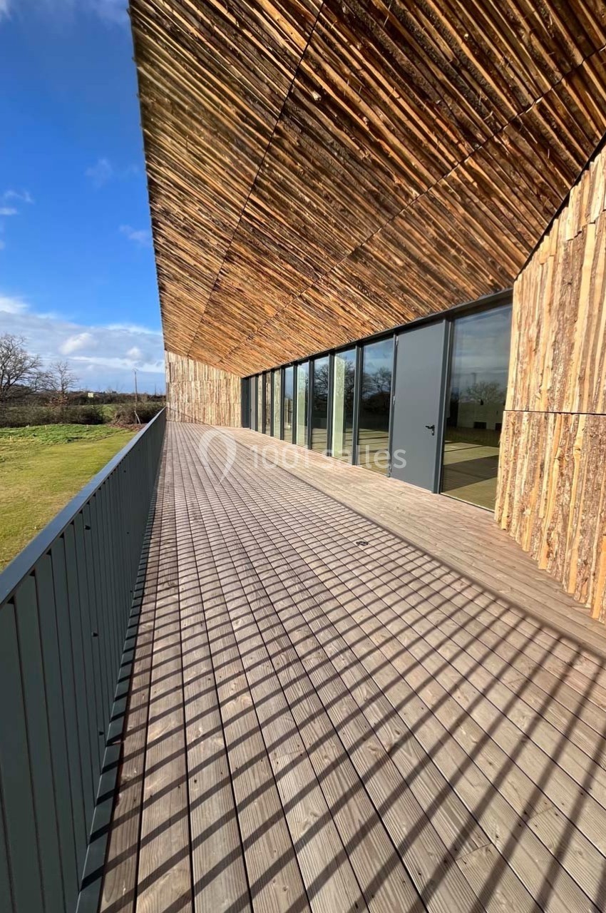 Façade en bois avec de grandes baies vitrées donnant sur une terrasse en bois, entourée d'un paysage verdoyant.