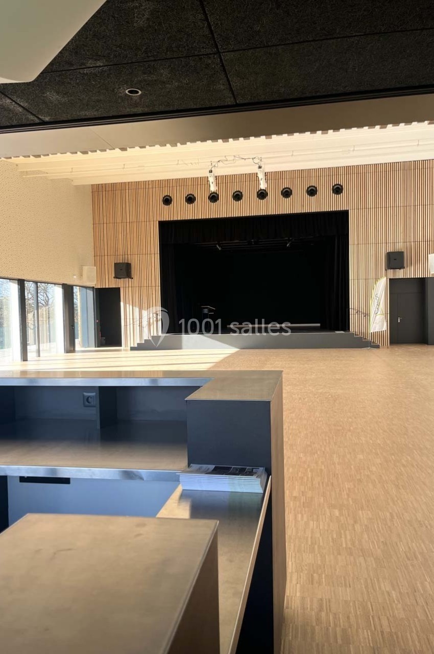 Salle polyvalente lumineuse avec scène surélevée, parquet clair, murs en bois et grandes baies vitrées.