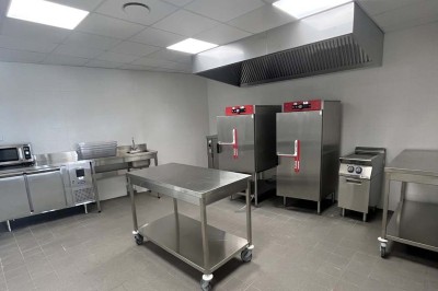 Cuisine professionnelle équipée avec plan de travail inox, fours, hotte, micro-ondes et sol carrelé gris.