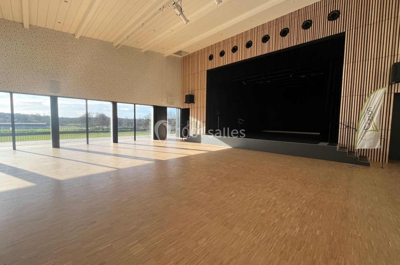 Salle polyvalente lumineuse avec parquet, grande scène noire et larges baies vitrées donnant sur un espace extérieur.