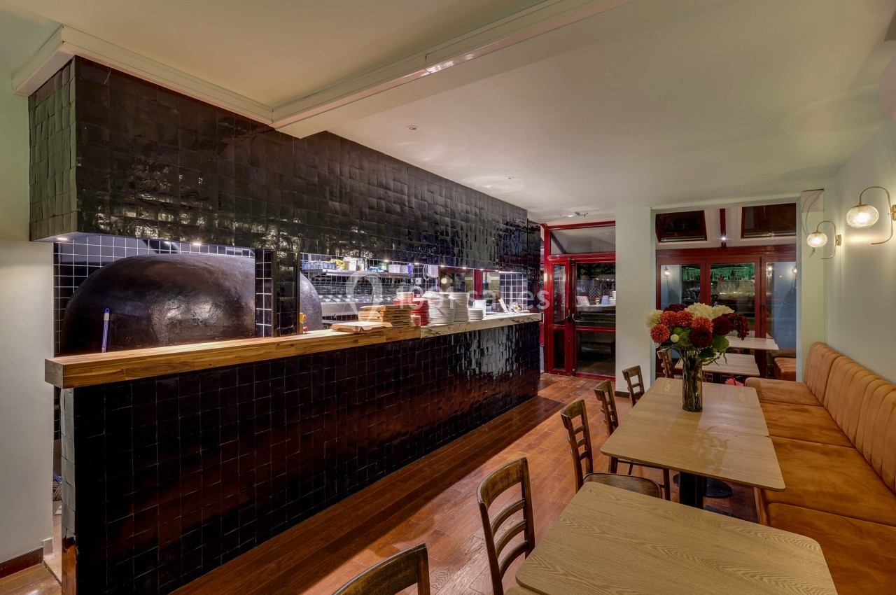 Intérieur d'un restaurant avec un comptoir en carrelage noir, un four à pizza et des tables en bois alignées.