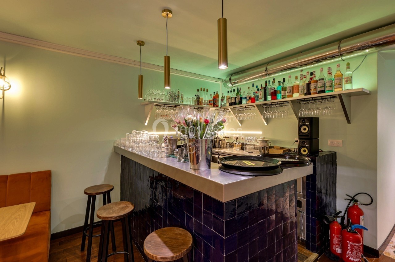 Bar avec comptoir en carrelage sombre, tabourets en bois, étagères avec bouteilles et verres, éclairage suspendu.