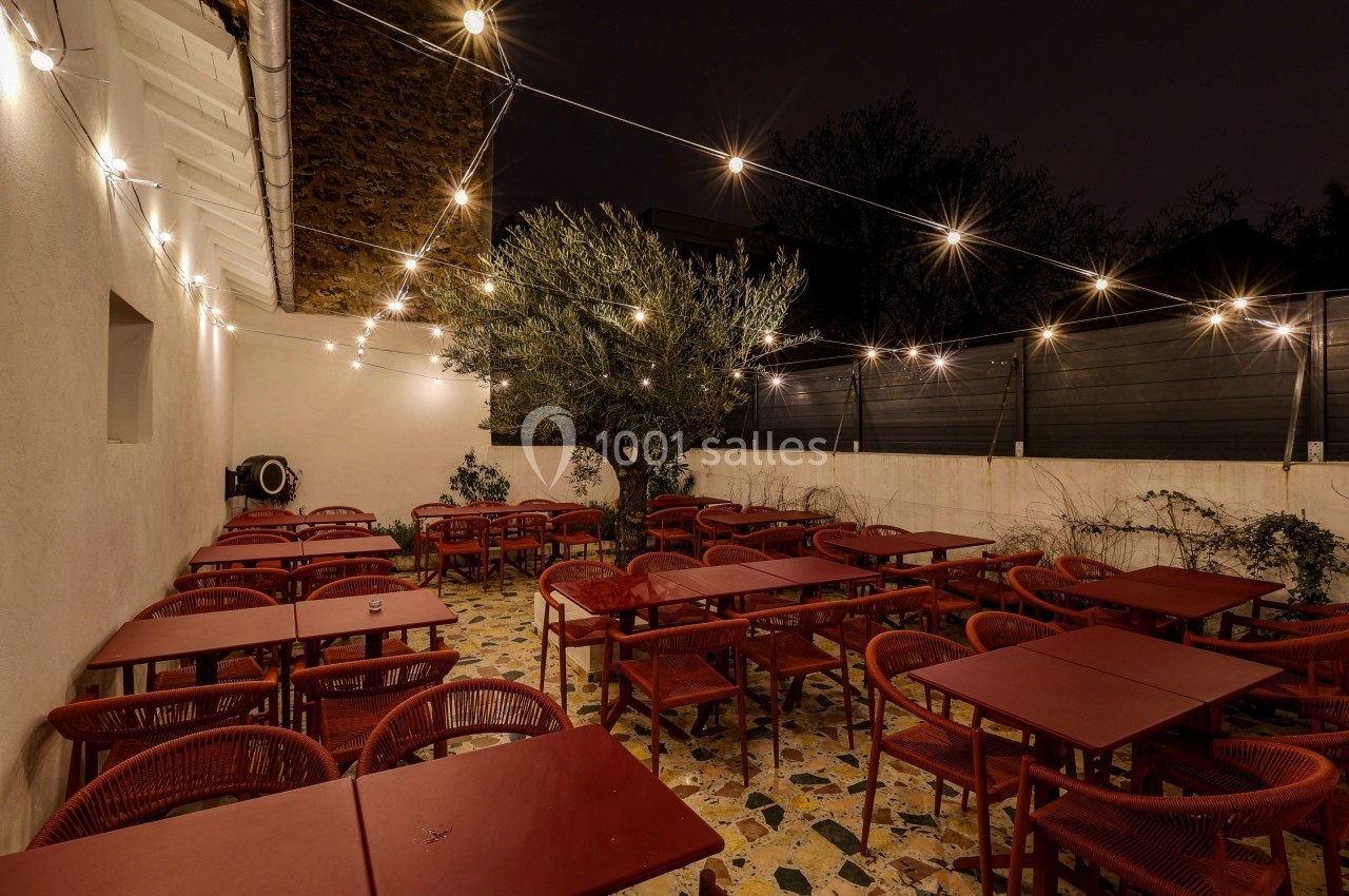 Terrasse extérieure avec tables et chaises rouges, éclairée par des guirlandes lumineuses sous un ciel nocturne.