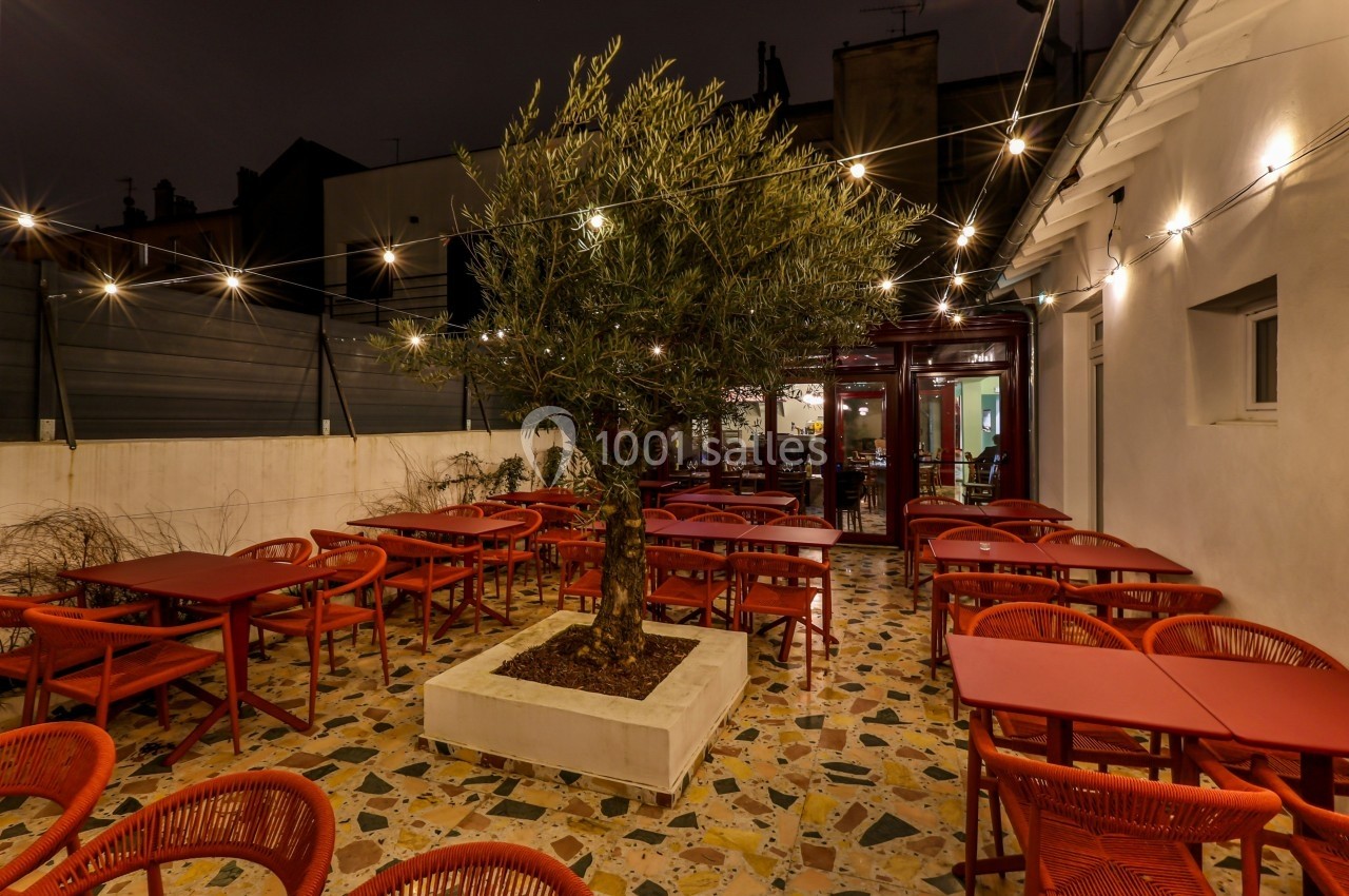 Terrasse extérieure d'un restaurant avec des tables rouges, un olivier central et des guirlandes lumineuses suspendues.