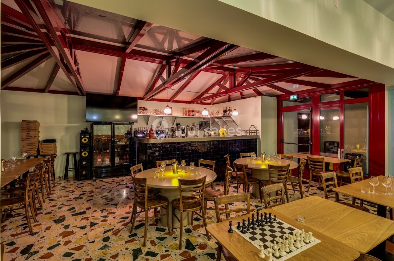 Salle de restaurant avec tables en bois, sol en terrazzo, bar éclairé et échiquier posé sur une table.