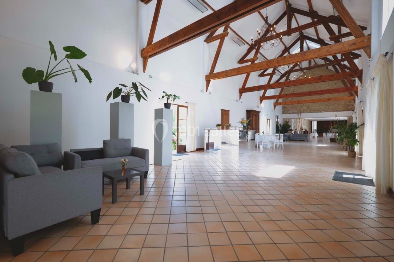 Grande salle lumineuse avec poutres apparentes, carrelage au sol, mobilier moderne et plantes décoratives.