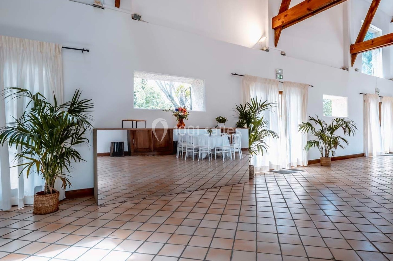 Salle lumineuse avec carrelage au sol, grandes fenêtres, plantes en pot et tables blanches dressées au fond.