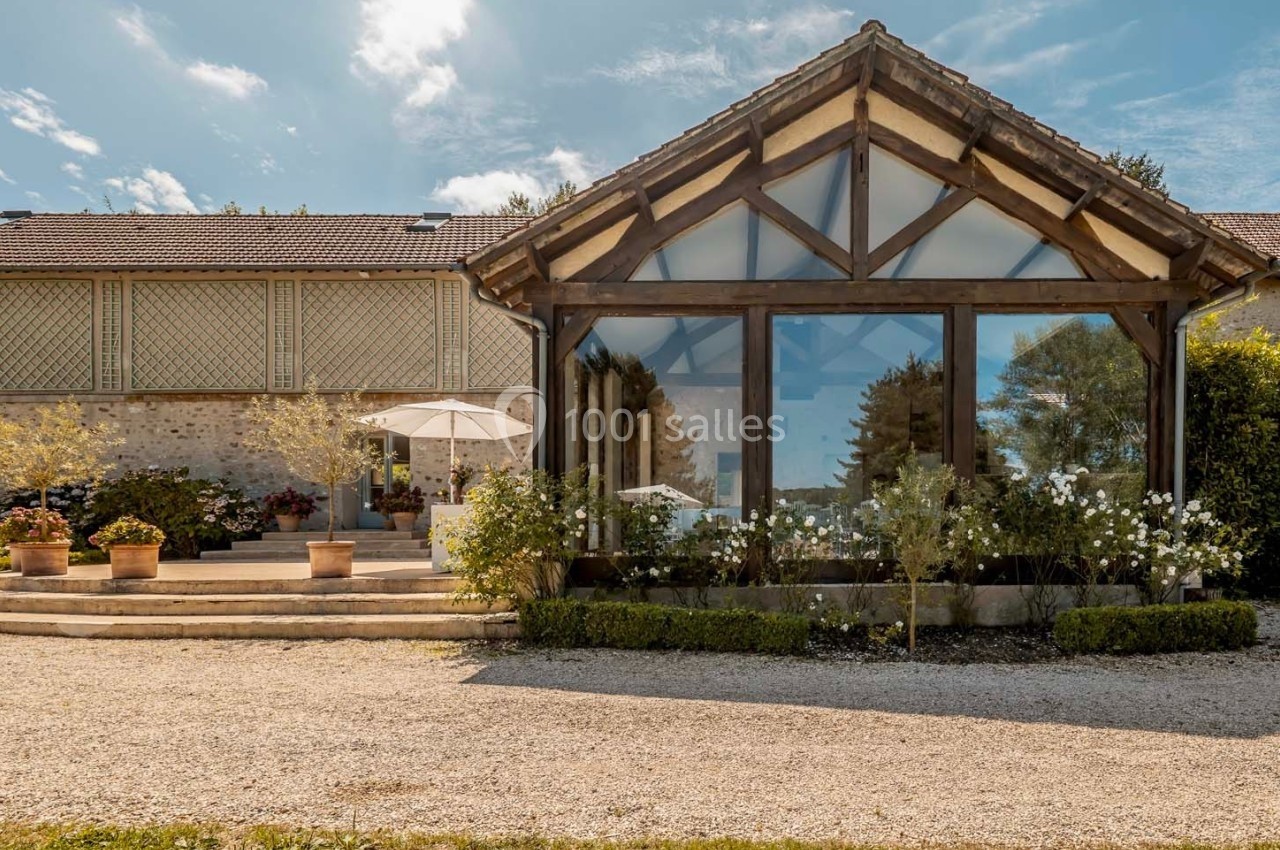 Façade d'un bâtiment avec une grande verrière, entourée de plantes et d'une terrasse avec parasol sous un ciel dégagé.