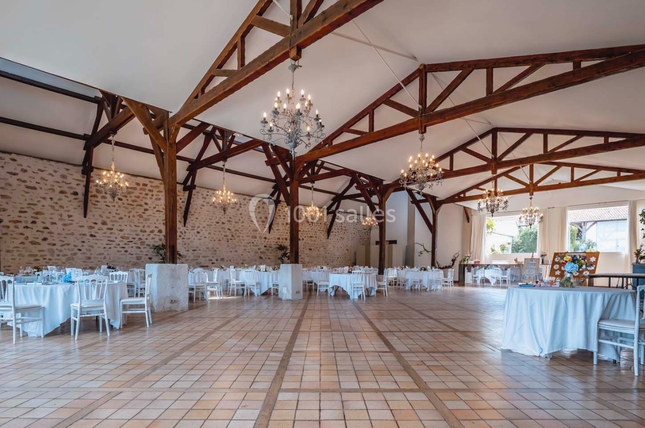 Salle spacieuse avec poutres apparentes, tables rondes dressées et lustres suspendus, sol en carrelage clair.