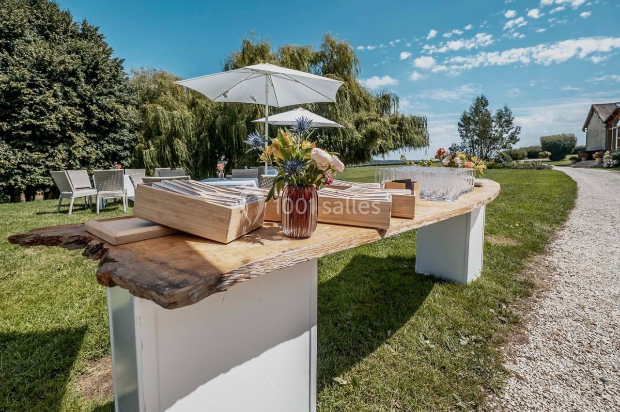 Table en bois brut avec des caisses en bois, des fleurs et des verres, installée dans un jardin sous des parasols.