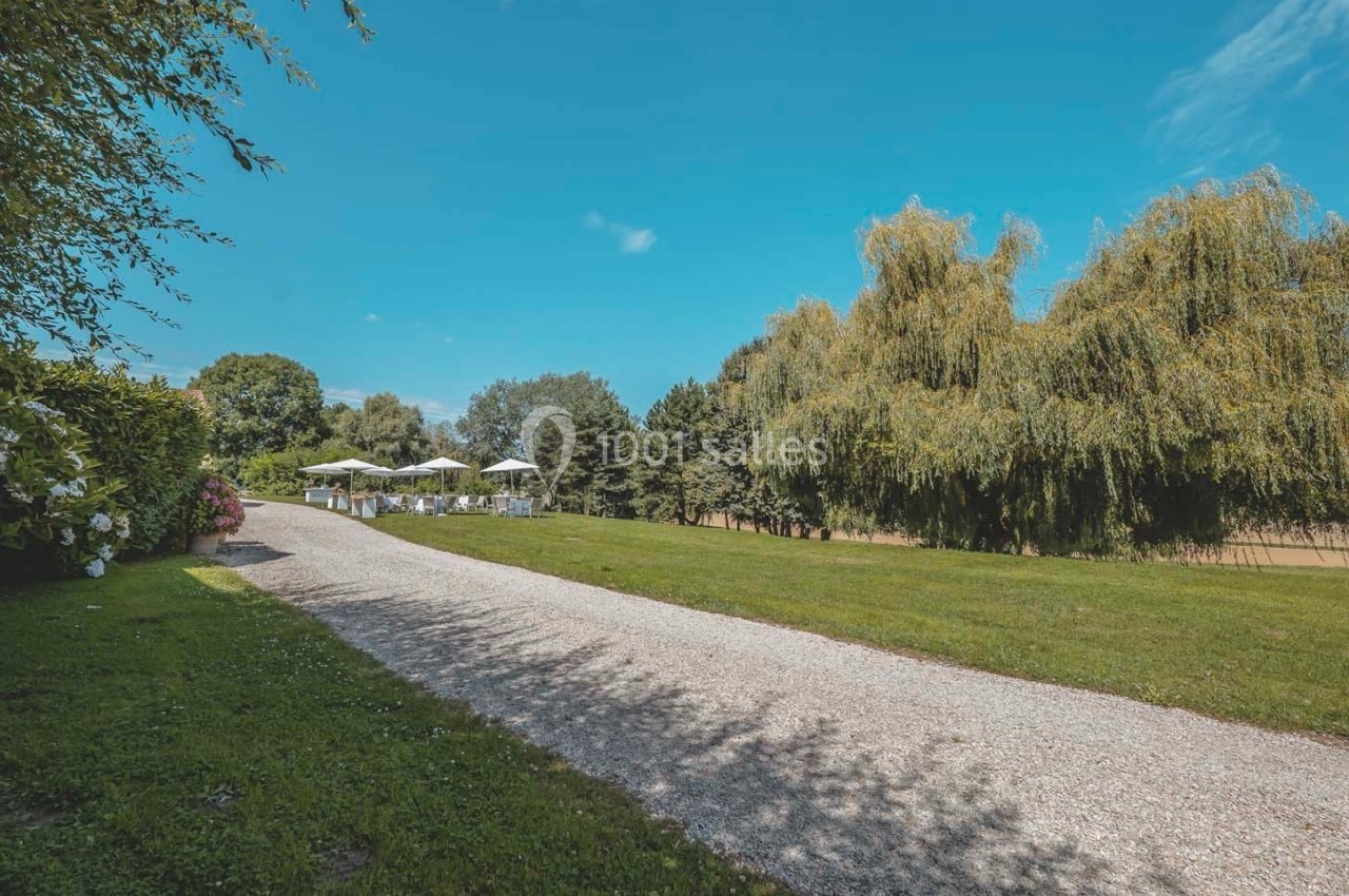 Allée de gravier bordée de pelouse et d'arbres, menant à des tables avec parasols sous un ciel bleu.