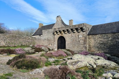 Vue aérienne d'un manoir en pierre entouré de champs et de végétation, avec un village et la mer à l'horizon.