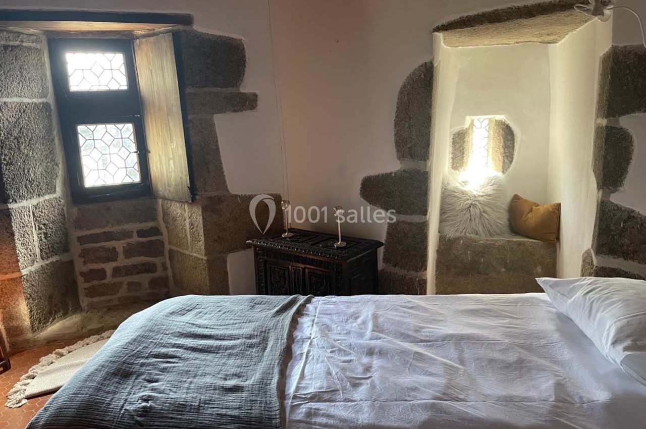 Chambre rustique avec lit simple, murs en pierre, fenêtre à vitraux et alcôve décorée de coussins.