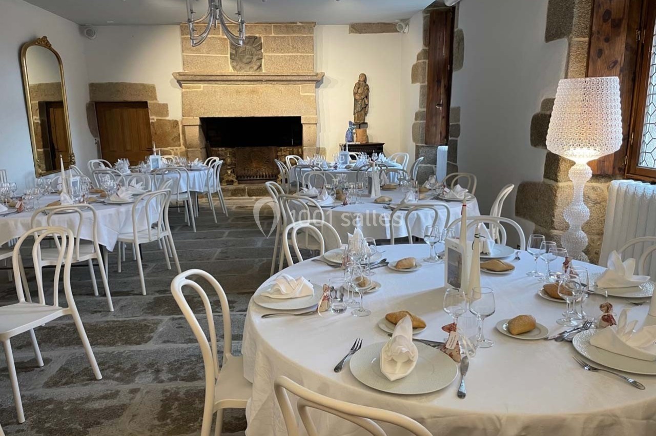 Salle de réception élégante avec tables rondes dressées, chaises blanches et cheminée en pierre.