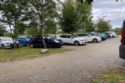 Vue aérienne d'un manoir en pierre entouré de champs et de végétation, avec un village et la mer à l'horizon.