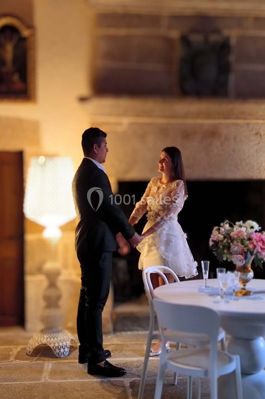 Un couple debout, se tenant les mains, dans une pièce chaleureuse avec une table décorée de fleurs au premier plan.