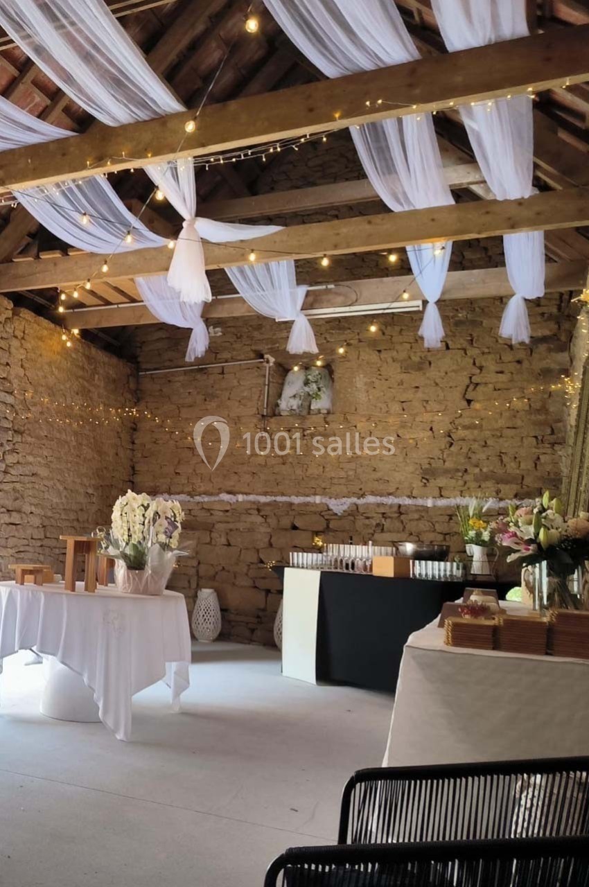 Salle en pierre avec plafond en poutres apparentes, décorée de voilages blancs, guirlandes lumineuses et tables dressées.