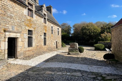 Vue aérienne d'un manoir en pierre entouré de champs et de végétation, avec un village et la mer à l'horizon.