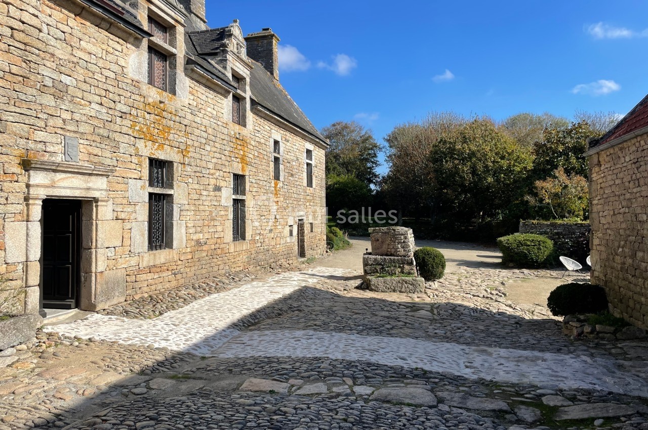 Cour pavée avec un bâtiment en pierre, un puits central et des arbres en arrière-plan sous un ciel bleu.