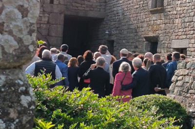 Vue aérienne d'un manoir en pierre entouré de champs et de végétation, avec un village et la mer à l'horizon.