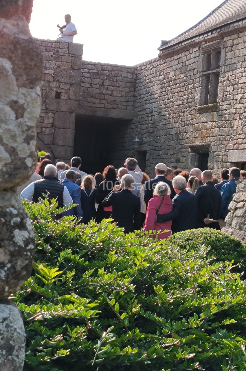 Un groupe de personnes assiste à une présentation en extérieur devant un bâtiment en pierre historique.